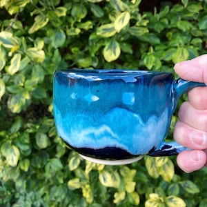 May include: A ceramic mug with a blue and white glaze. The mug has a rounded shape with a handle and a white base. The glaze transitions from dark blue to light blue and white, creating a unique, artistic appearance. The mug is handmade.