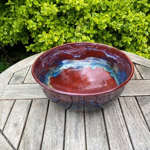 May include: A handcrafted ceramic bowl with a glossy finish. The bowl has a deep red interior with blue and gold accents around the rim. The exterior has a textured, dark red appearance. The bowl sits on a weathered wooden table.