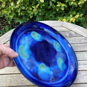 May include: Oval, deep blue ceramic platter with a glossy finish. The platter has a dark blue centre with lighter blue and green accents. The rim is a darker shade of blue. The platter is on a wooden surface.