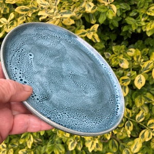 Puede incluir: Un plato de cerámica ovalado hecho a mano con un esmalte turquesa texturizado y moteado y un borde plateado. El plato se sostiene en una mano, con un fondo de follaje verde y amarillo.