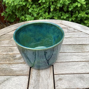 May include: A round ceramic bowl featuring a glossy glaze in shades of deep teal and forest green. The exterior surface displays subtle, dark vertical line patterns that contrast with the smooth, blended colour transitions of the finish.