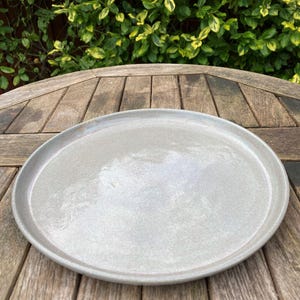 May include: A round, pale grey ceramic plate with a slightly raised edge. The plate is on a wooden table. The plate is approximately 30 cm in diameter, suitable for serving appetizers or desserts.