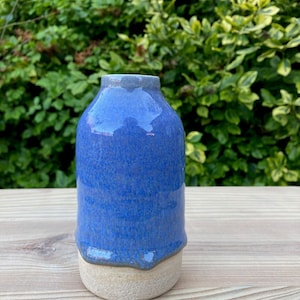 Könnte beinhalten: Eine kleine, blaue Keramikvase mit weißem Sockel. Die Vase hat eine strukturierte, gesprenkelte Glasur und einen leicht unregelmäßigen Rand.