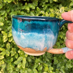 May include: A ceramic mug featuring a glossy glaze that transitions from a deep dark blue at the rim to a lighter turquoise and white in the centre, ending in a natural tan clay base. The mug has a curved handle and a wide, rounded body shape.