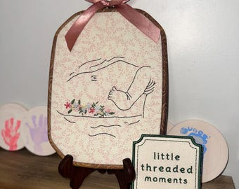 May include: An oval embroidery hoop frame featuring a cream fabric with a pink floral pattern. The embroidery depicts a black line art outline of a pregnant belly with a small foot and colorful flowers. A pink satin ribbon bow sits at the top.