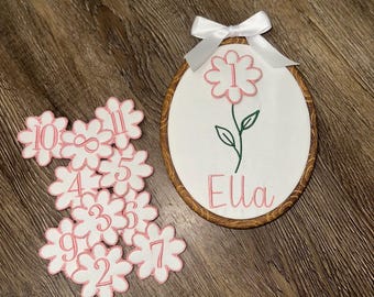 May include: An oval wooden embroidery hoop containing white fabric with a pink embroidered flower, the number 1, and the name Ella, topped with a white ribbon bow. Beside it are several white flower-shaped fabric cutouts with pink numbers.