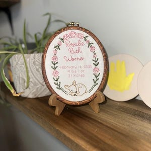May include: An oval embroidery hoop with a faux wood grain frame, displaying white fabric with hand-stitched pink flowers, green vines, and a fawn. Text reads: Rosalie Ruth Worner, February 16, 2025, 9 lbs 7 oz, 21 inches. It rests on a small wooden stand.
