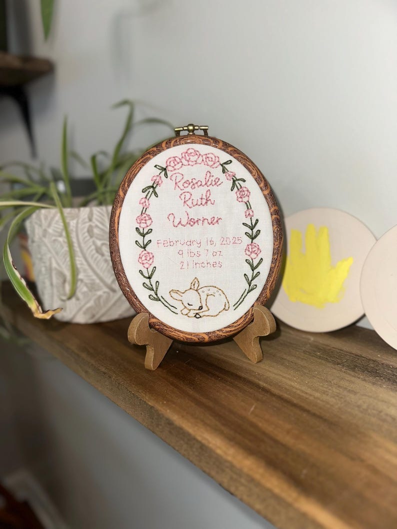 May include: An oval embroidery hoop with a faux wood grain frame, displaying white fabric with hand-stitched pink flowers, green vines, and a fawn. Text reads: Rosalie Ruth Worner, February 16, 2025, 9 lbs 7 oz, 21 inches. It rests on a small wooden stand.
