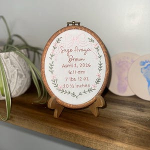May include: An oval wooden embroidery hoop containing white fabric with hand-stitched text reading Sage Avaya Brown, April 2, 2026, 6:11 am, 7 lbs 12 oz, and 20 1/2 inches, framed by an embroidered pink bow and green leafy vine border.