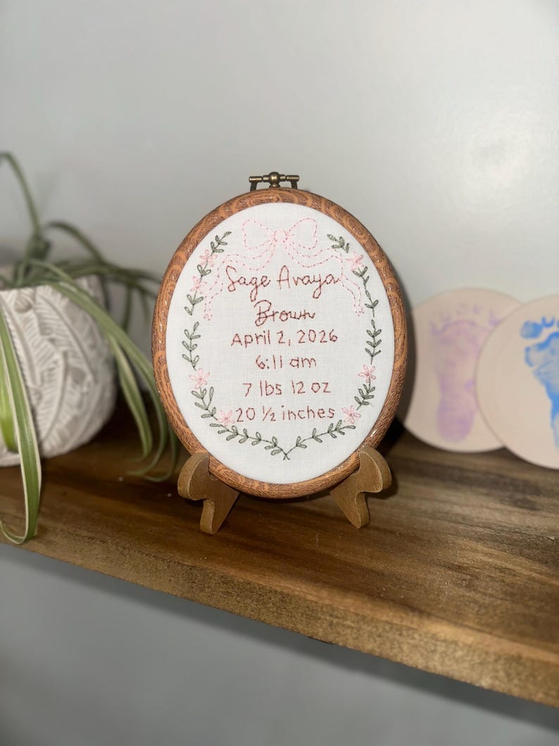 May include: An oval wooden embroidery hoop containing white fabric with hand-stitched text reading Sage Avaya Brown, April 2, 2026, 6:11 am, 7 lbs 12 oz, and 20 1/2 inches, framed by an embroidered pink bow and green leafy vine border.