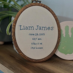 May include: An oval embroidery hoop with a faux wood grain frame containing off-white fabric. The text is embroidered in dark blue thread: Liam James, June 20, 2025, 6:27 am, 6 lbs 11 oz, and 19.5 inches. The hoop has a metal tightening screw at the top.
