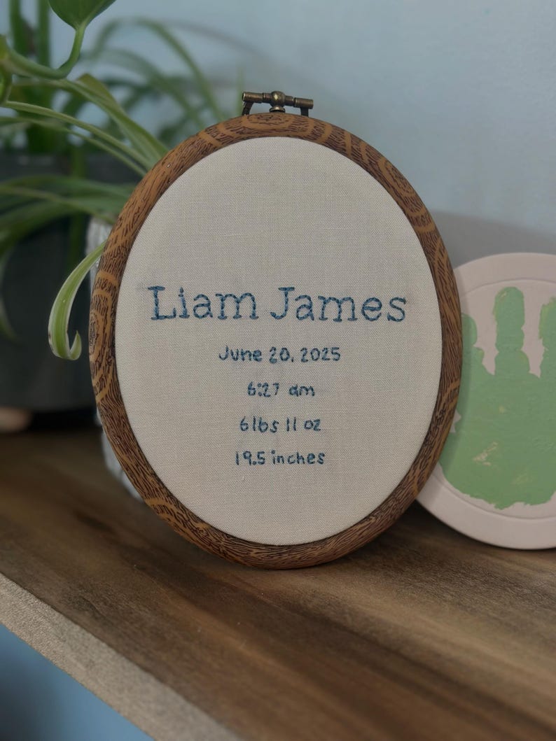 May include: An oval embroidery hoop with a faux wood grain frame containing off-white fabric. The text is embroidered in dark blue thread: Liam James, June 20, 2025, 6:27 am, 6 lbs 11 oz, and 19.5 inches. The hoop has a metal tightening screw at the top.