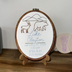 May include: An oval embroidery hoop with a wood-grain frame featuring a stitched mountain landscape, a deer, and a bear. The text reads: Luke Clayton, January 24, 2024, 7:33 pm, 8 lbs 13 oz, 21.5 inches. The hoop rests on a small wooden display stand.