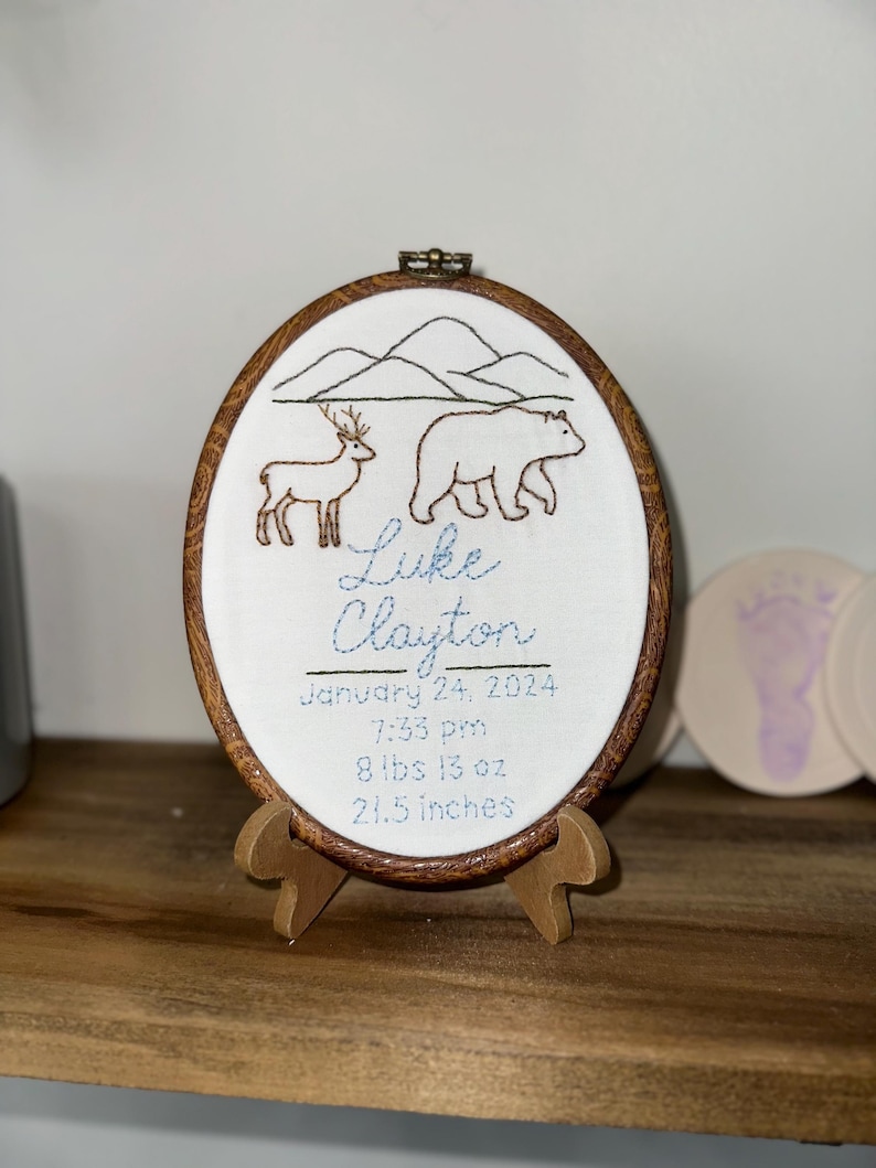 May include: An oval embroidery hoop with a wood-grain frame featuring a stitched mountain landscape, a deer, and a bear. The text reads: Luke Clayton, January 24, 2024, 7:33 pm, 8 lbs 13 oz, 21.5 inches. The hoop rests on a small wooden display stand.