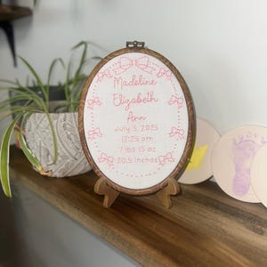 May include: An oval embroidery hoop containing white fabric with pink stitched text and bow motifs. The text reads: Madeline Elizabeth Ann, July 3, 2025, 12:25 pm, 7 lbs 15 oz, 20.5 inches. The hoop is displayed on a small wooden stand.