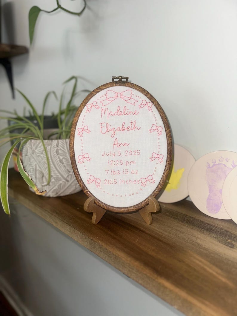 May include: An oval embroidery hoop containing white fabric with pink stitched text and bow motifs. The text reads: Madeline Elizabeth Ann, July 3, 2025, 12:25 pm, 7 lbs 15 oz, 20.5 inches. The hoop is displayed on a small wooden stand.