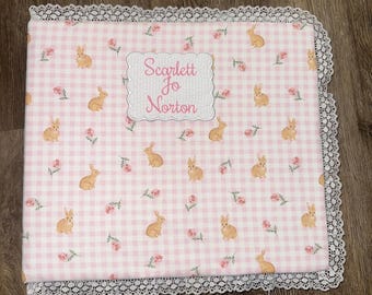 May include: A rectangular baby blanket featuring a pink and white gingham pattern with small brown rabbits and pink flowers. The blanket has a white lace trim border and a central white scalloped label with the text Scarlett Jo Norton.