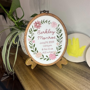 May include: An oval wooden embroidery hoop containing white fabric with floral stitching and text reading Berkley Monroe, July 29, 2025, 11:26 pm, 6 lbs 12 oz, 19.5 inches. The hoop is displayed on a small wooden stand with a dark brown finish.