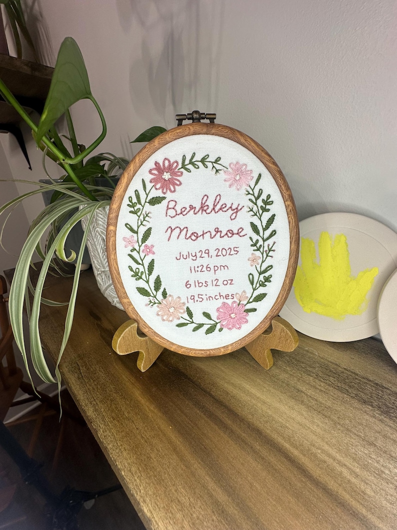 May include: An oval wooden embroidery hoop containing white fabric with floral stitching and text reading Berkley Monroe, July 29, 2025, 11:26 pm, 6 lbs 12 oz, 19.5 inches. The hoop is displayed on a small wooden stand with a dark brown finish.