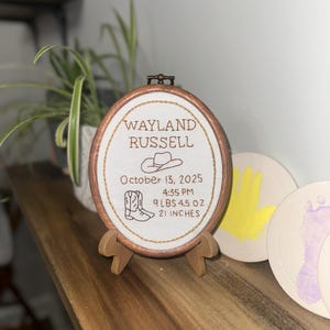 May include: An oval wooden embroidery hoop containing white fabric with brown stitched text reading Wayland Russell, October 13, 2025, 4:35 PM, 9 lbs 4.5 oz, 21 inches, featuring an embroidered cowboy hat and a pair of cowboy boots.