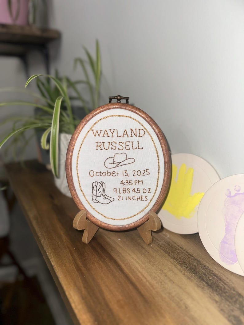 May include: An oval wooden embroidery hoop containing white fabric with brown stitched text reading Wayland Russell, October 13, 2025, 4:35 PM, 9 lbs 4.5 oz, 21 inches, featuring an embroidered cowboy hat and a pair of cowboy boots.