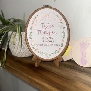 May include: An oval embroidery hoop containing white fabric with hand-stitched text reading Tylee Morgan, 7LBS 2OZ, 19 INCHES, and DECEMBER 27, 2024. The design features a stitched peach-colored bow at the top and a green floral wreath border.