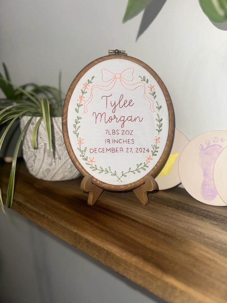 May include: An oval embroidery hoop containing white fabric with hand-stitched text reading Tylee Morgan, 7LBS 2OZ, 19 INCHES, and DECEMBER 27, 2024. The design features a stitched peach-colored bow at the top and a green floral wreath border.