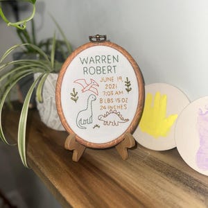 May include: An oval wooden embroidery hoop containing white fabric with stitched text reading WARREN ROBERT, JUNE 19, 2021, 7:03 AM, 8 LBS 15 OZ, 24 INCHES. The design features embroidered green and red dinosaurs and foliage on a small wooden stand.