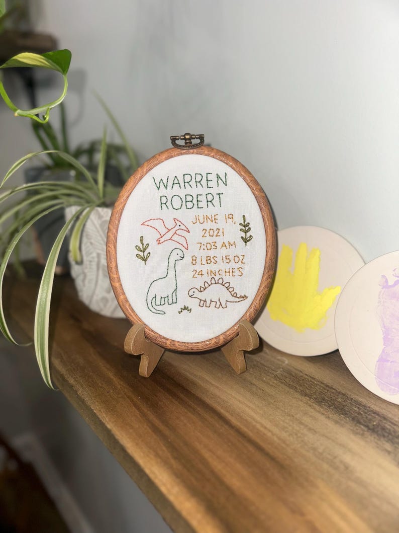 May include: An oval wooden embroidery hoop containing white fabric with stitched text reading WARREN ROBERT, JUNE 19, 2021, 7:03 AM, 8 LBS 15 OZ, 24 INCHES. The design features embroidered green and red dinosaurs and foliage on a small wooden stand.
