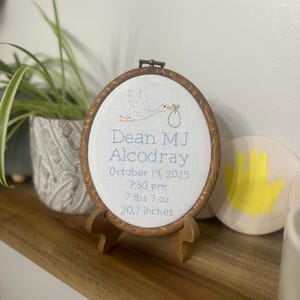 May include: An oval embroidery hoop with a wood-grain frame containing white fabric. It features a stitched stork carrying a bundle and text: Dean MJ Alcodray, October 19, 2025, 7:30 pm, 7 lbs 7 oz, 20.7 inches. The hoop rests on a small wooden stand.
