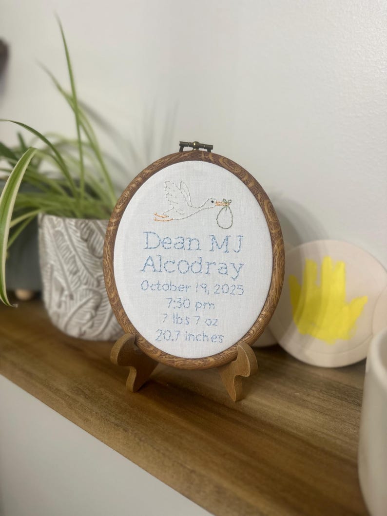 May include: An oval embroidery hoop with a wood-grain frame containing white fabric. It features a stitched stork carrying a bundle and text: Dean MJ Alcodray, October 19, 2025, 7:30 pm, 7 lbs 7 oz, 20.7 inches. The hoop rests on a small wooden stand.