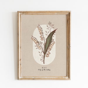 May include: Framed botanical print of Lily of the Valley. The artwork has a beige background with an oval shape, and the text "MAY" and "Lily of the Valley". The frame is a distressed, light brown colour.