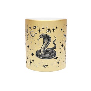 Könnte beinhalten: Ein goldfarbener, metallisch glänzender Becher mit einer zentralen schwarzen Illustration einer zusammengerollten Kobra. Das Design ist von verschiedenen kleinen schwarzen Sternen und dünneren Schlangenumrissen umgeben.