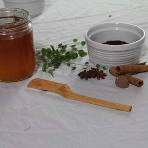 Puede incluir: Un tarro de vidrio lleno de miel, un tazón de cerámica blanco con café molido, palitos de canela, anís estrellado y una cuchara de madera sobre un mantel blanco.