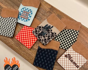 May include: A collection of leatherette phone wallets with various designs. Patterns include a black and white lightning bolt, red checkerboard, a cactus with the word "Howdy", a black and white checkerboard, a navy blue pattern, and a tan and white checkerboard with the word "Mama".