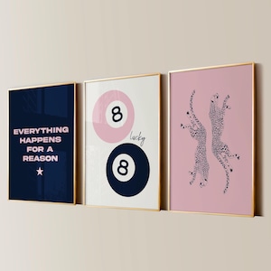 Op de afbeelding: Drie ingelijste kunstdrukken met gouden frames. Eén print heeft de tekst "EVERYTHING HAPPENS FOR A REASON" op een marineblauwe achtergrond. De tweede print heeft een roze en marineblauw 8-ball ontwerp met het woord "lucky". De derde print toont twee cheeta's op een roze achtergrond.