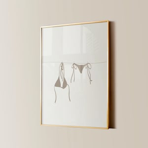 May include: A framed print of a minimalist line drawing of a bikini top and bottom hanging on a clothesline. The print is on a white background and is framed in a gold frame.