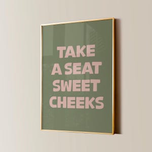 May include: A framed print with a green background and pink text that reads "Take a seat sweet cheeks".