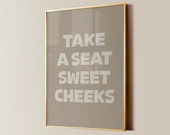 May include: A gold framed print with a brown background and white text that reads "Take a seat sweet cheeks".