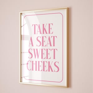 Puede incluir: Una impresión enmarcada en dorado con el texto "TAKE A SEAT SWEET CHEEKS" en letras rosas sobre fondo blanco.