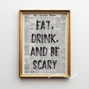 May include: A framed print with a gold-colored frame. The print features a vintage newspaper background with the words "EAT. DRINK. AND BE SCARY" in bold, black letters. The print is a decorative item.