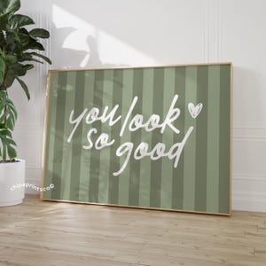 May include: A framed art print with a green striped background and the words "you look so good" in white script. A small white heart is next to the word "good". The frame is a light brown color.