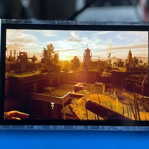 May include: A white handheld gaming device with gold buttons and a screen displaying a video game. The game features a sunset over a city with buildings and trees.