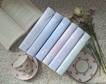 May include: A stack of six blue and purple paperback books with a floral design and the text "The Secret Garden" on the cover. The books are on a floral patterned surface with a cup and saucer and a bouquet of flowers.