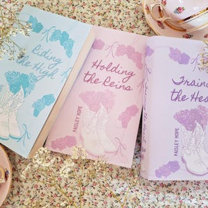 May include: Three books with floral and cowboy boot illustrations. The book covers are light blue, pink, and lavender, with titles "Riding the High", "Holding the Reins", and "Training the Heart". The author's name is "Paisley Hope".
