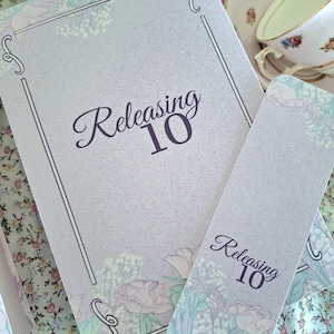 Puede incluir: Un diario y un marcapáginas de color lila claro con ilustraciones florales y el texto "Releasing 10". El diario tiene un borde decorativo y el marcapáginas es de forma rectangular. Una taza y un platillo están en el fondo.