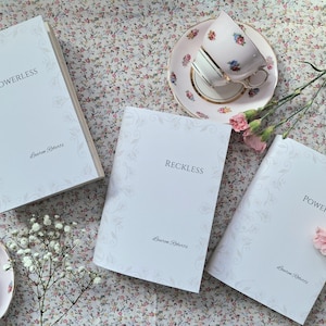 Puede incluir: Tres libros blancos con bordes florales decorativos y los títulos "Powerless", "Reckless" y "Powerful" sobre una superficie con estampado floral. Cada libro tiene el nombre del autor "Lauren Roberts" impreso en la portada.