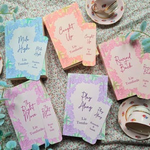May include: A collection of books and bookmarks with floral designs and titles like "Mile High," "Caught Up," and "Rewind It Back." The books are in pastel colors, and teacups and floral accents are also present.