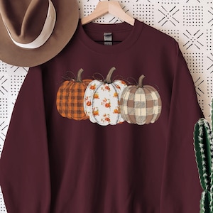 May include: A maroon sweatshirt featuring three pumpkins with different patterns: orange plaid, white floral, and tan plaid. A brown hat hangs to the left, and a cactus in a white pot is on the right. The sweatshirt is hanging on a wooden hanger.
