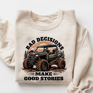 May include: Cream-colored sweatshirt featuring a graphic of a black off-road vehicle with large tires, set against a sunset and trees. The text "BAD DECISIONS MAKE GOOD STORIES" is arched above and below the vehicle.